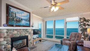 A living room with a stone fireplace, a television, and a large window overlooking the ocean. - Promo! Top Floor, Corner Oceanfront Condo - Indoor Pool/Hot Tub/Wifi and HDTV! (Depoe Bay)