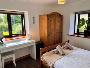 A bedroom with a desk, a bed with towels, a wooden wardrobe, and a view of greenery through the windows.