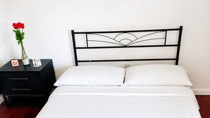 Uma cama com roupa de cama branca, cabeceira de metal preto e mesa de cabeceira de madeira com um vaso de rosas vermelhas.