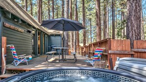 A hot tub area with two lounge chairs and an umbrella, surrounded by wooden deck and forest backdrop. - Larch Chalet- 1BR, 1BA 2 Min to Heavenly, Hot Tub (South Lake Tahoe)