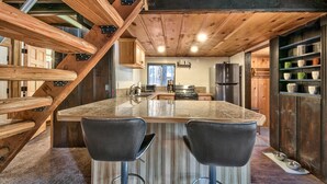 A kitchen with a large island, two bar stools, wooden beams, and a built-in shelf with dishes. - Larch Chalet- 1BR, 1BA 2 Min to Heavenly, Hot Tub (South Lake Tahoe)