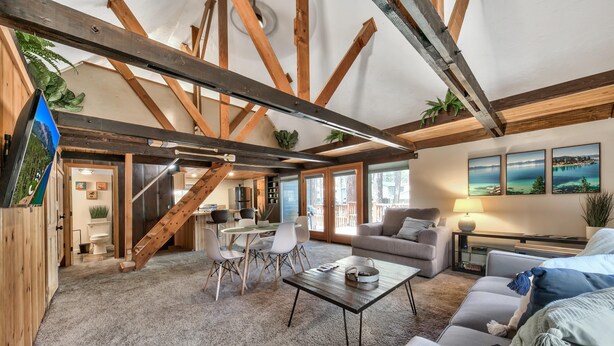 A spacious living area with a high ceiling, exposed wooden beams, a dining area with modern chairs, a sofa, and a coffee table. - Larch Chalet- 1BR, 1BA 2 Min to Heavenly, Hot Tub (South Lake Tahoe)