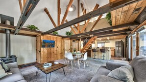 A spacious living area with a high ceiling, wooden beams, a fireplace, a dining table with chairs, and a kitchen in the background. - Larch Chalet- 1BR, 1BA 2 Min to Heavenly, Hot Tub (South Lake Tahoe)