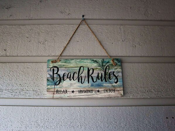 A wooden sign with the text "Beach Rules" and the words "RELAX," "UNWIND," and "ENJOY" hanging on a wall. - Sun, Sand and Pool : Condo at Savannah, Tybee (Tybee Island)