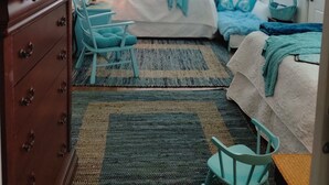 A room with a wooden dresser, two beds with blue bedding, a chair, a lamp, and a window with blinds. - Sunkissed Cottage (Gulfport)