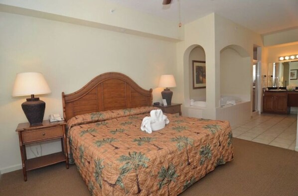 A hotel room with a wooden bed, two bedside tables with lamps, a bathroom with a mirror and lights, and a hallway leading to another room.