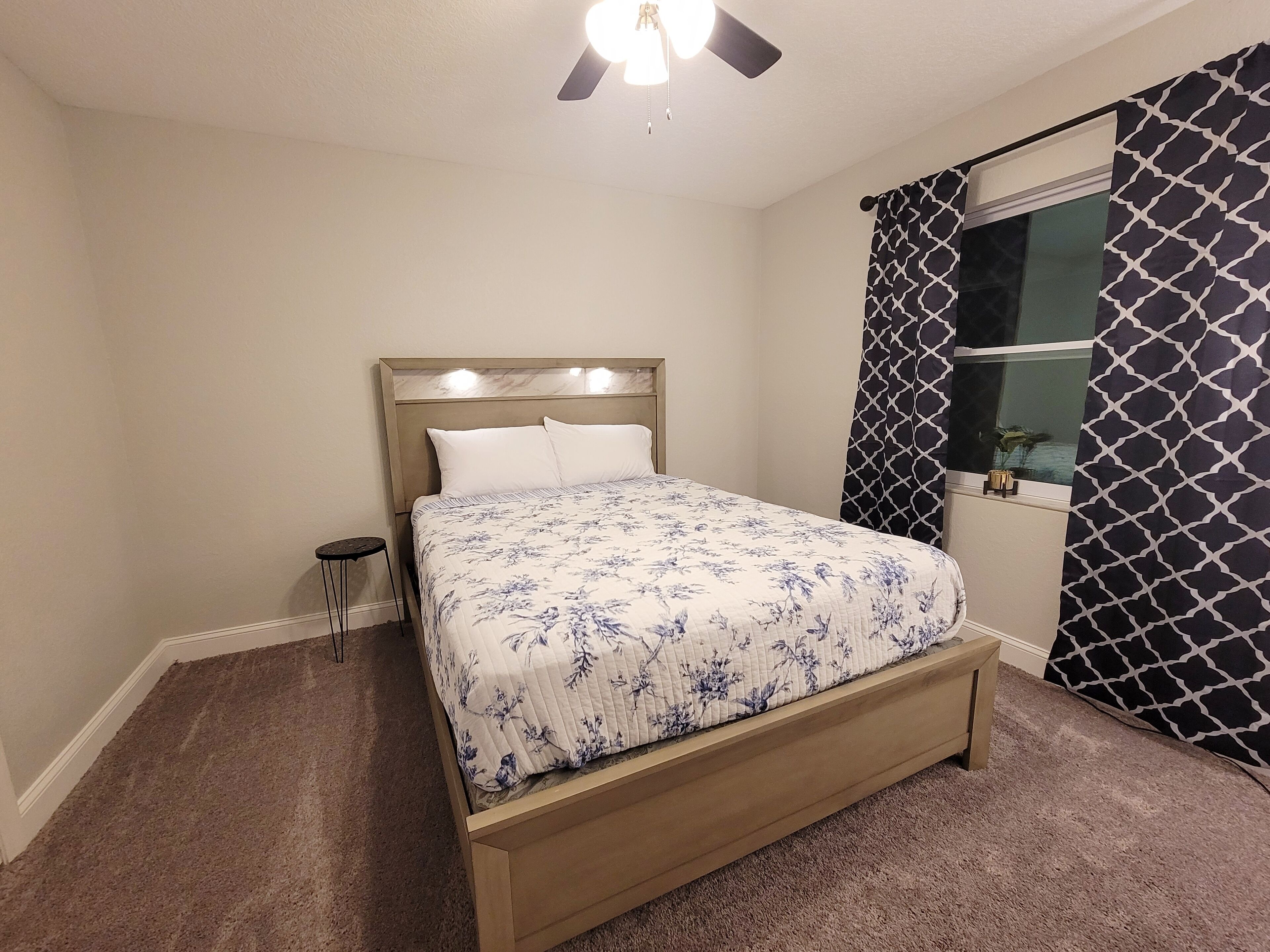 A bedroom with a bed, a small stool, a ceiling fan, and a window with patterned curtains.