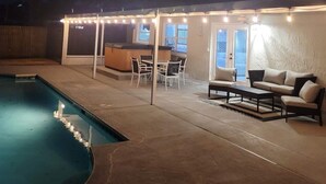 Un patio trasero con piscina, un porche techado con luces colgantes y mobiliario exterior.