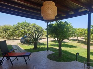 Un patio couvert doté d’un luminaire au plafond fait de fibres tressées, un coin salon avec une chaise et une table, et une vue sur un vignoble en arrière-plan.
