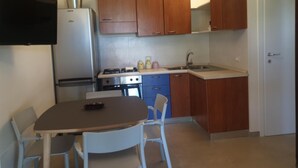 Une cuisine compacte équipée d’un réfrigérateur, d’une cuisinière, d’un évier et de meubles de rangement en bois. On y trouve une table avec deux chaises et un téléviseur fixé au mur.