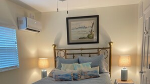 Ein Schlafzimmer mit einem Bett, zwei Nachttischen mit Lampen, einem Deckenventilator, einem Fenster mit Jalousien und einem gerahmten Bild an der Wand.