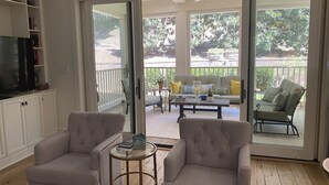Ein Wohnzimmer mit Fernseher, zwei Sesseln, einem Couchtisch und einer Schiebetür zu einer Terrasse mit einer Couch und Stühlen.