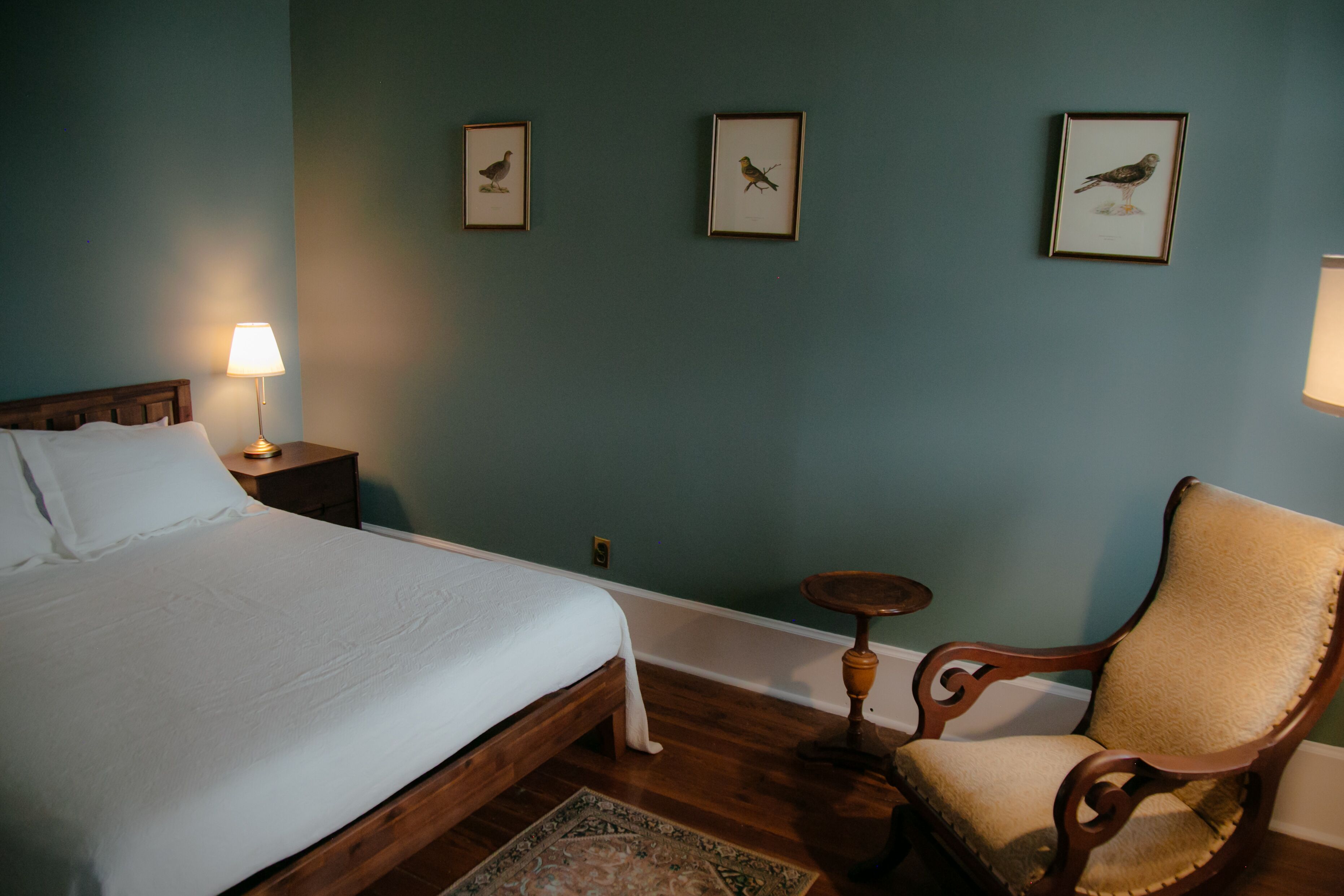 A bedroom with a bed, a nightstand with a lamp, a chair, and framed pictures on the wall.