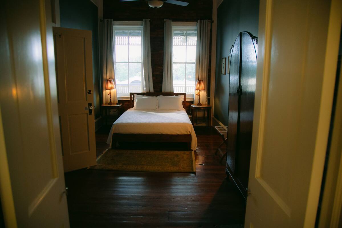 Une chambre à coucher comprenant un lit, deux lampes de chevet, un ventilateur de plafond et une vue sur l’extérieur grâce à deux fenêtres.