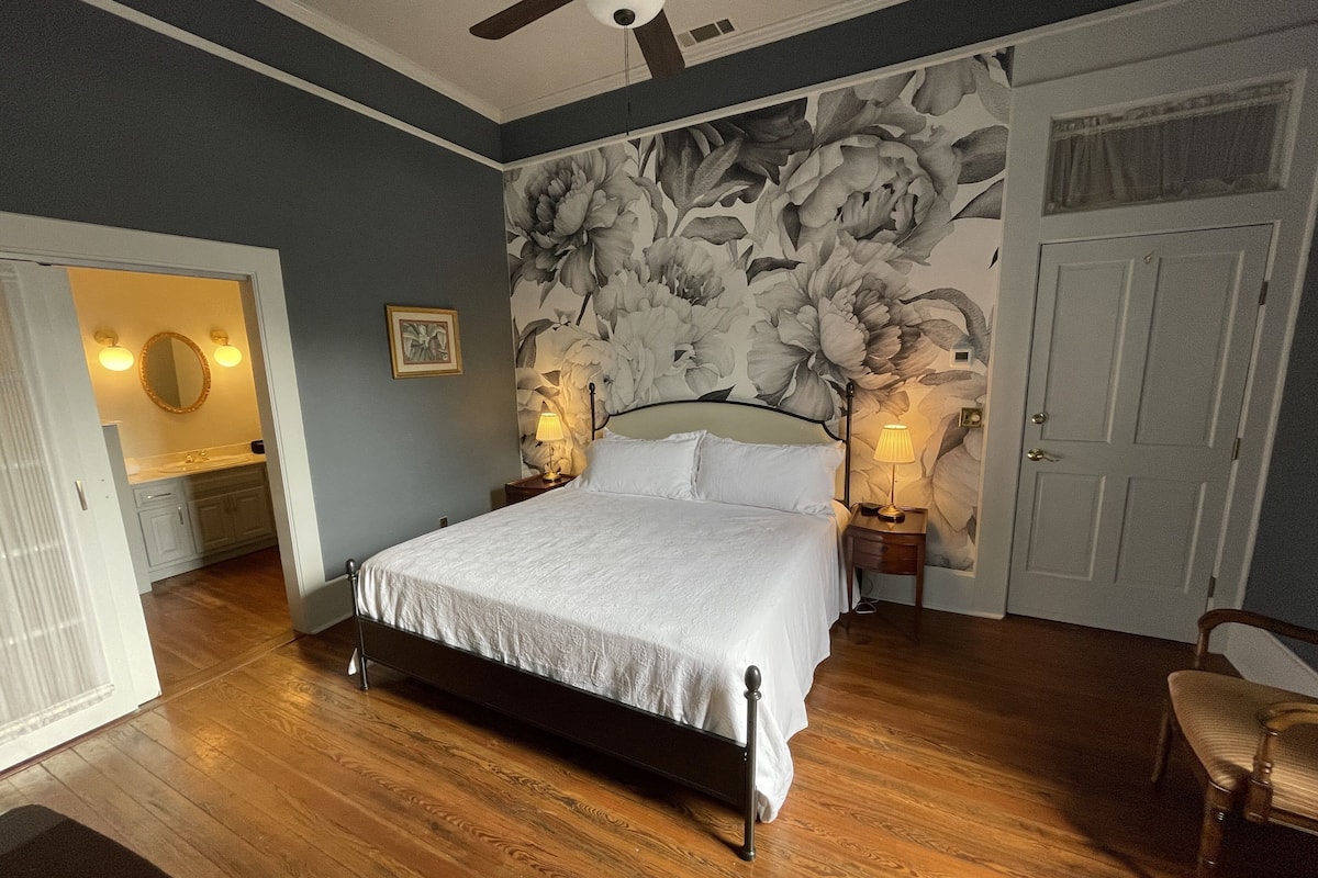 Une chambre à coucher avec un grand lit, deux lampes de chevet, un ventilateur de plafond, un papier peint à motifs floraux et un plancher en bois.