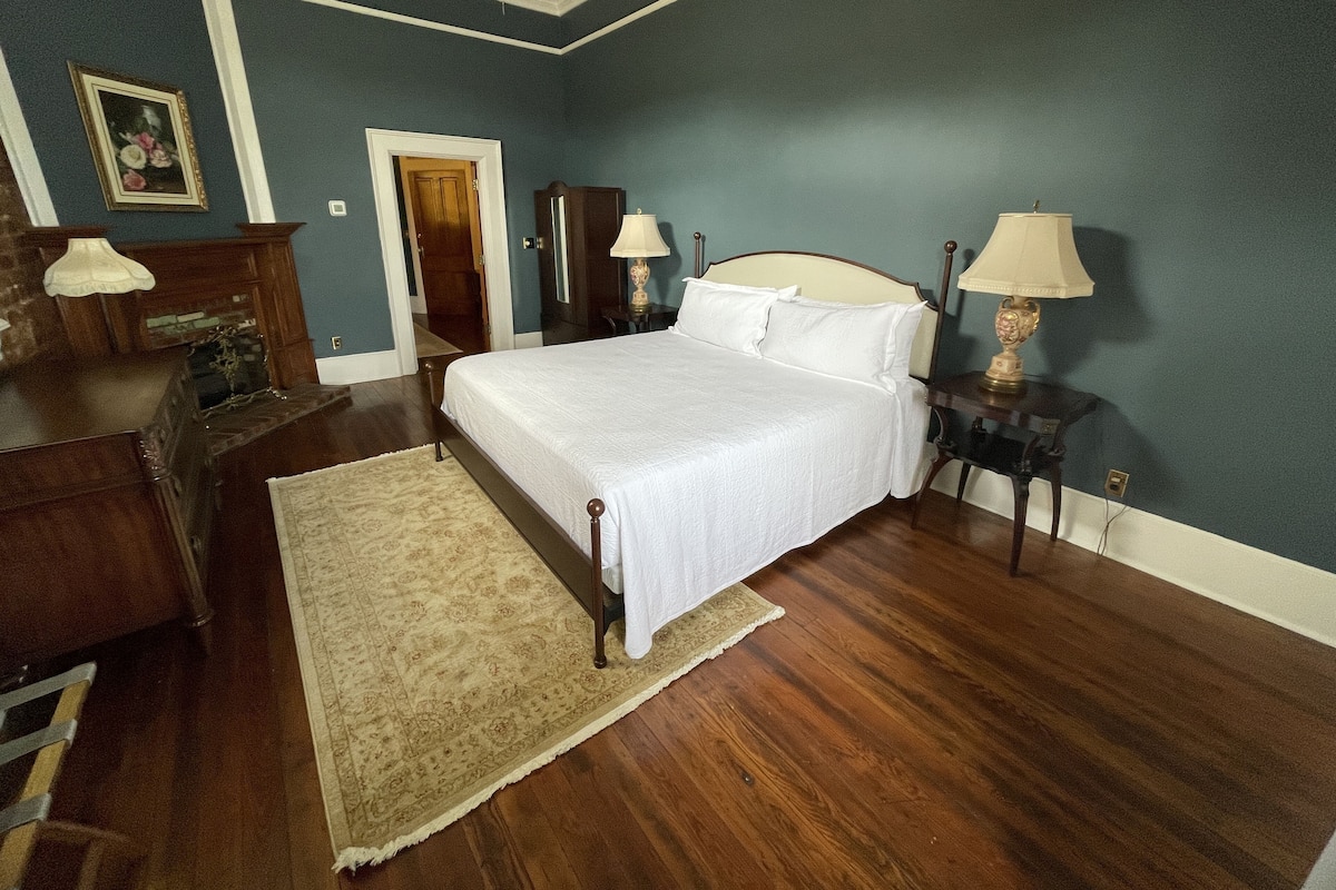 Une chambre à coucher comprenant un lit, une commode en bois, deux tables de chevet avec des lampes, une porte et un tapis à motifs.