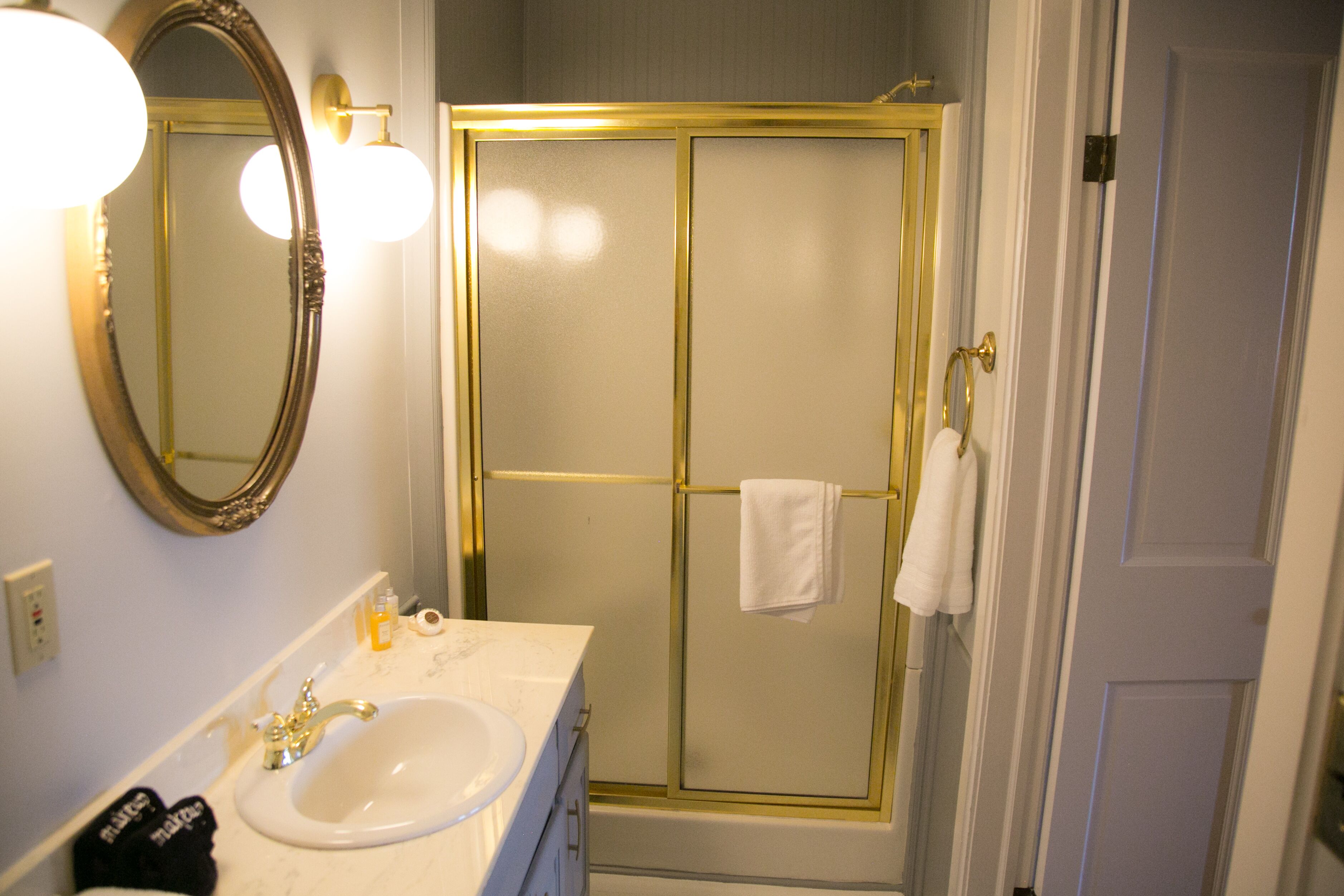 Un baño con lavamanos blanco, espejo con marco dorado y cabina de ducha con paredes de vidrio.