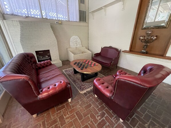 A room with a brick floor, a white brick wall, a red leather sofa, a red leather armchair, a wooden coffee table with a chessboard, a white armchair, and a window with white curtains. - The Grand Hotel (Hogansville)