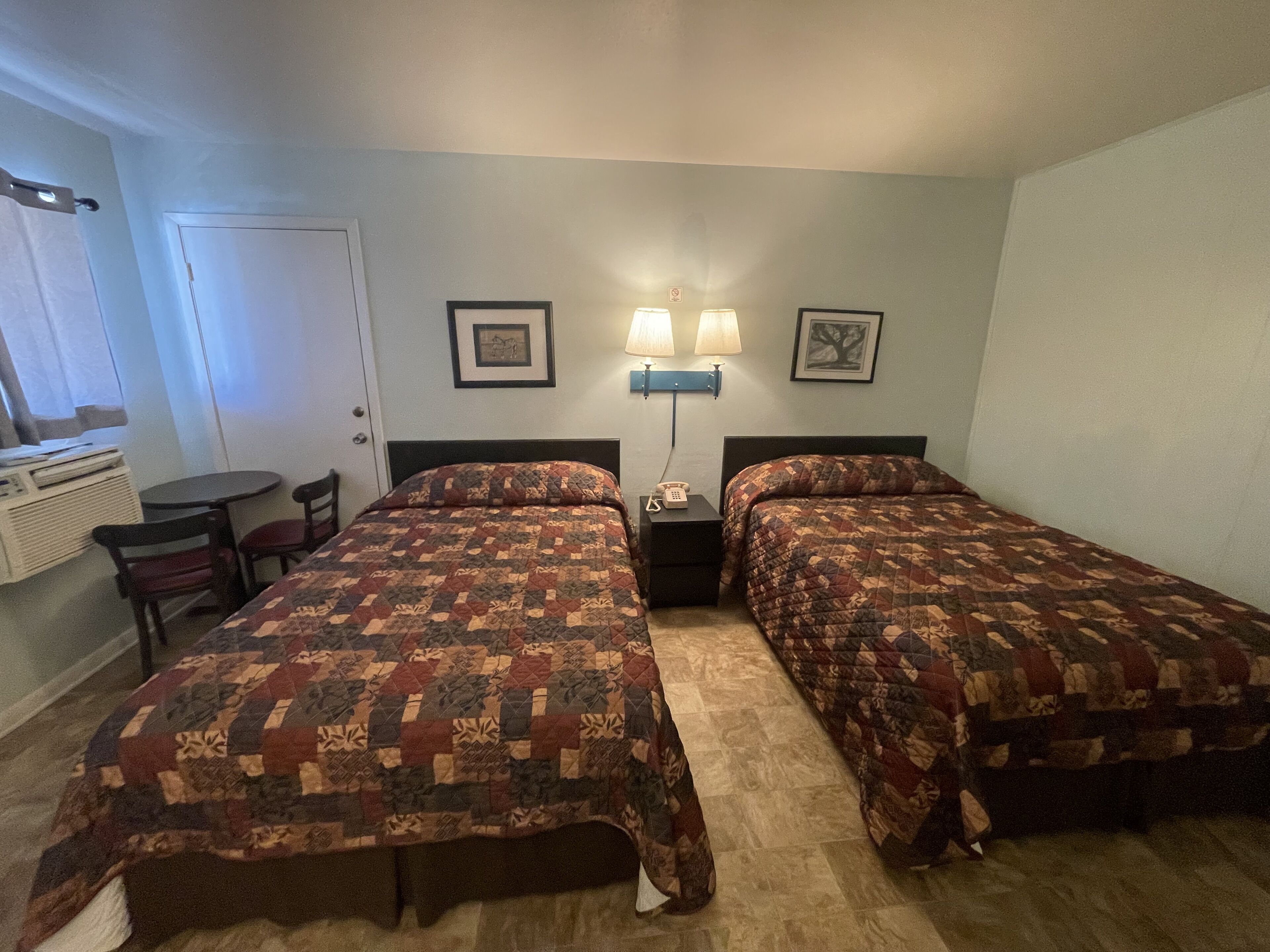 A hotel room with two beds, a small table, chairs, a desk, and a lamp.