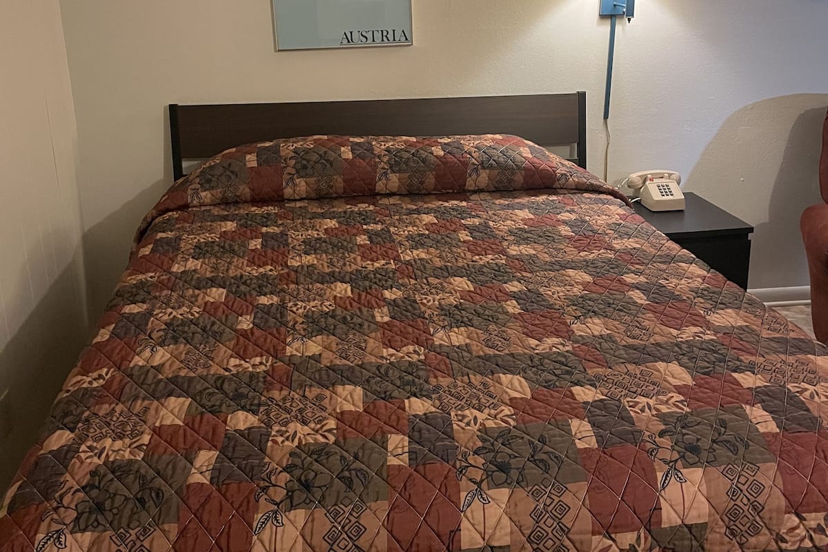 Ett hotellrum med en säng, ett nattduksbord med en lampa, en inramad bild av Österrike och en telefon.