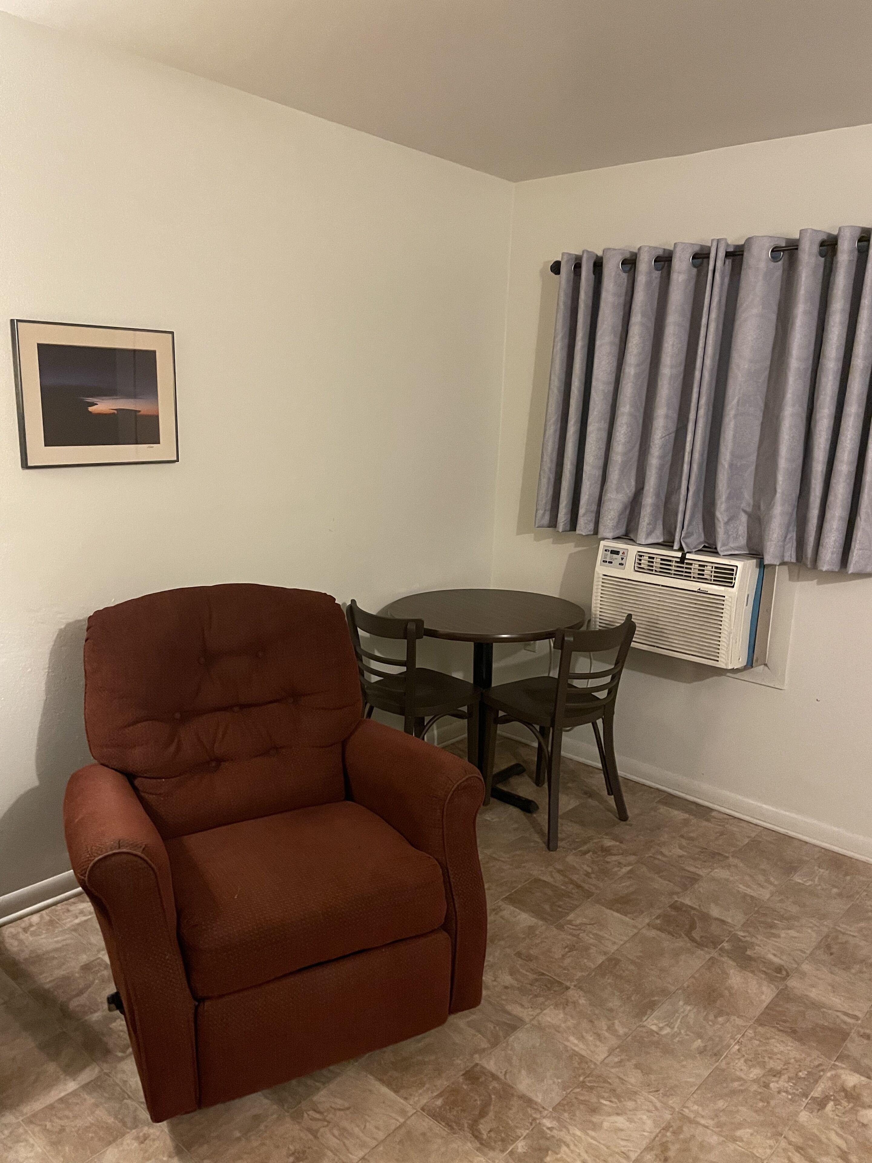 A room with a brown armchair, a small round table with chairs, a picture on the wall, and a window with curtains.
