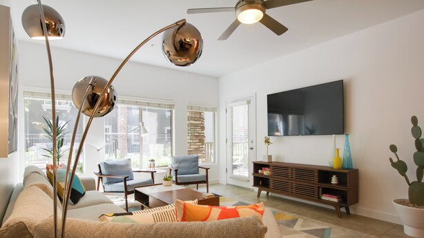 A modern living room with a sofa, armchairs, a coffee table, and a TV mounted on the wall. There is a ceiling fan, a floor lamp, and a bookshelf with decorative items.