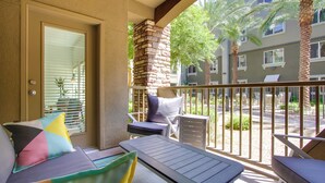 A balcony with a sofa, a small table, and a chair. There are palm trees and a building in the background.
