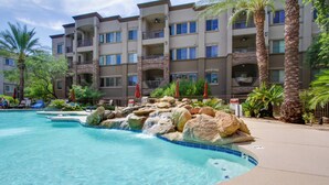 A hotel with a swimming pool featuring a rock waterfall, palm trees, and outdoor seating.