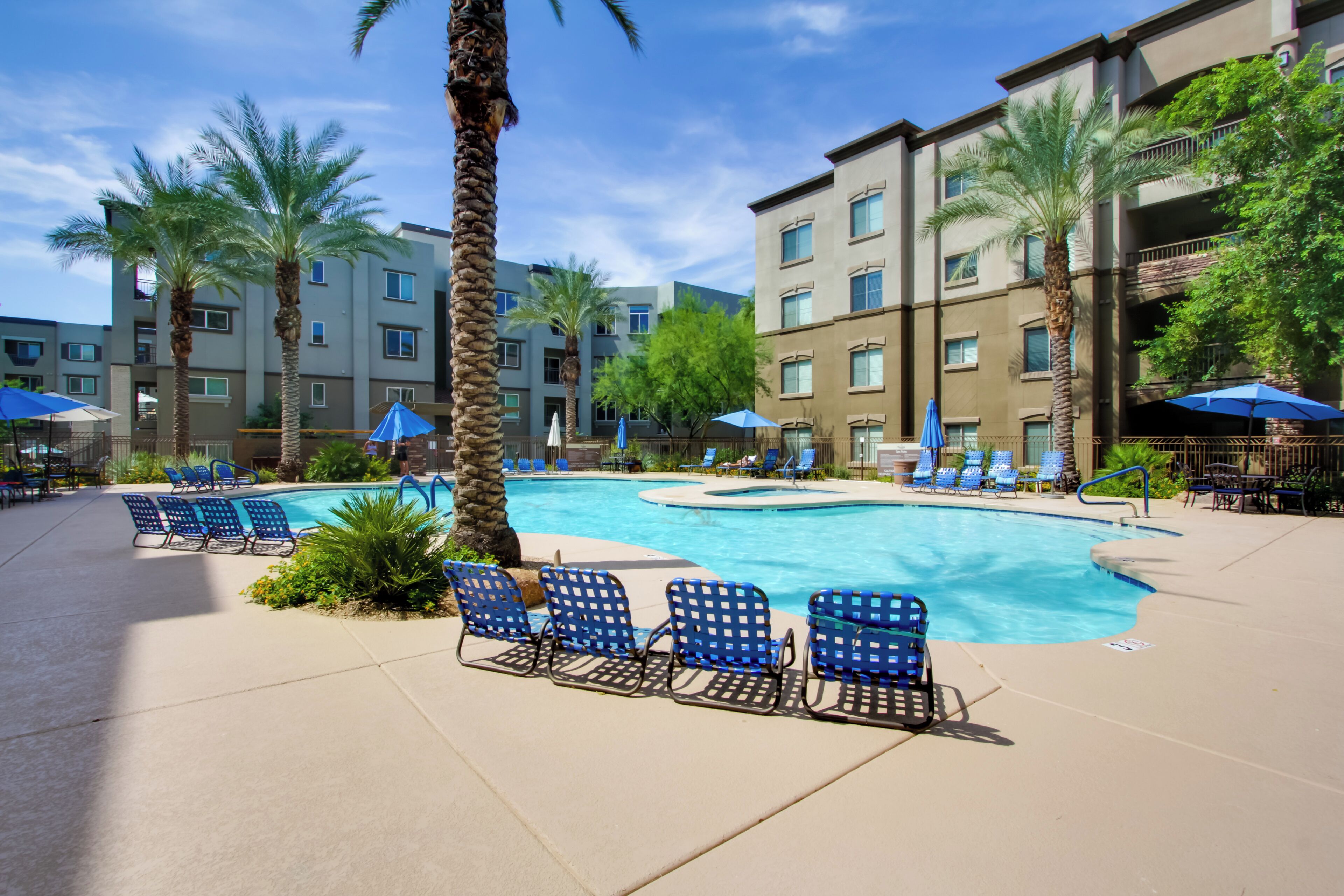 A hotel pool area with blue chairs and umbrellas, surrounded by palm trees and a multi-story building.