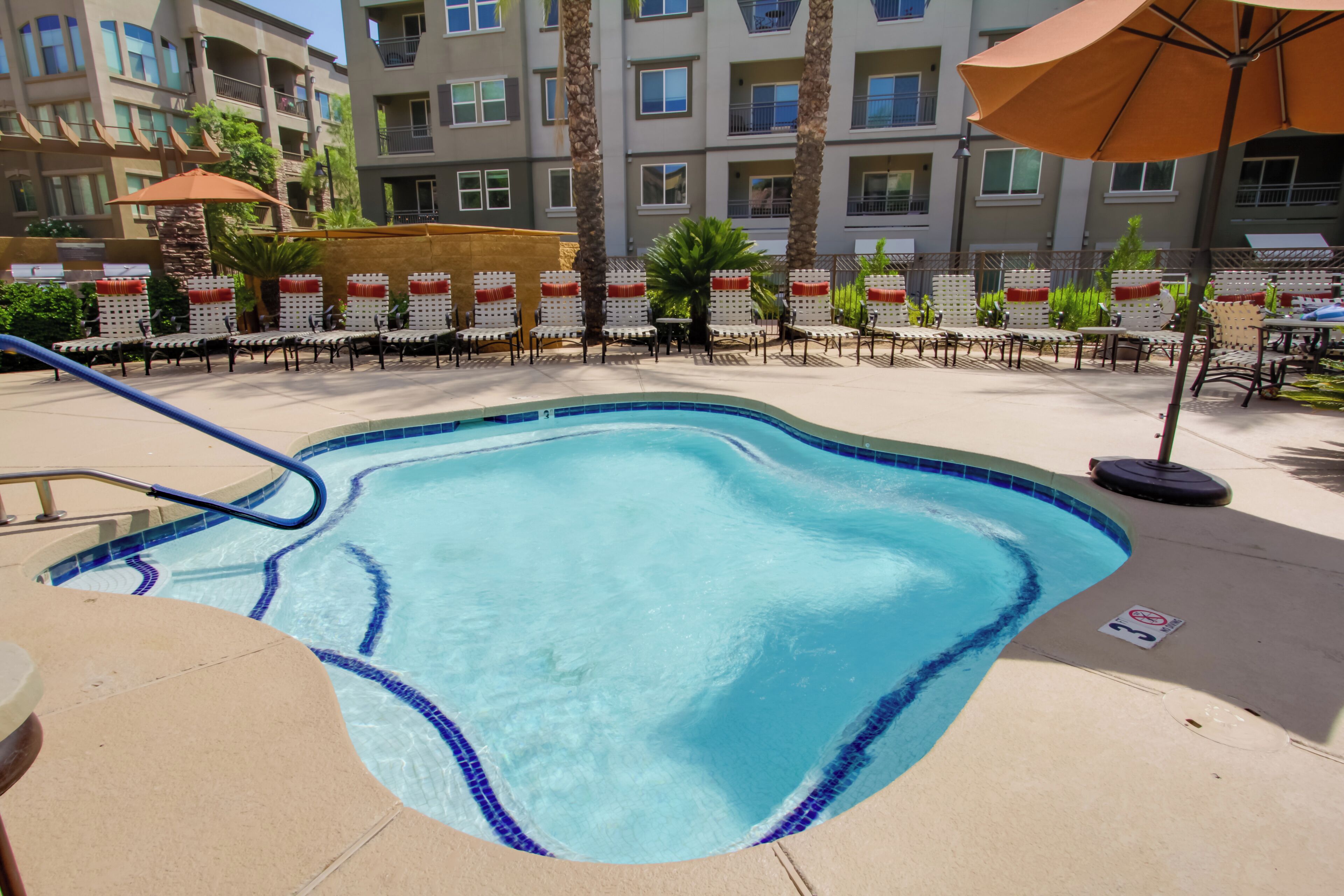 A swimming pool with lounge chairs and umbrellas, surrounded by a fence and a building with balconies.