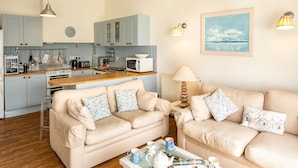 A modern living room with a beige sofa set, a glass coffee table, and a kitchen area with light blue cabinets and a wooden countertop. - Slipway 12 (Wadebridge)