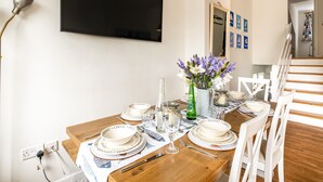 A dining area with a wooden table set for four, white plates with text, a vase with flowers, and a TV mounted on the wall. - Slipway 12 (Wadebridge)