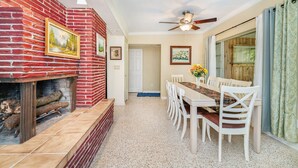 Un comedor con chimenea de ladrillo, sillas blancas, una mesa larga y un ventilador de techo.