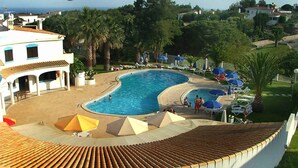 Una piscina con tumbonas y sombrillas, rodeada de palmeras y un edificio con techo de tejas.