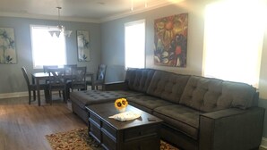 Ein Wohnzimmer mit Ecksofa, Couchtisch mit einer Vase voller Sonnenblumen, Teppich mit Muster und Essbereich mit Stühlen und Tisch.