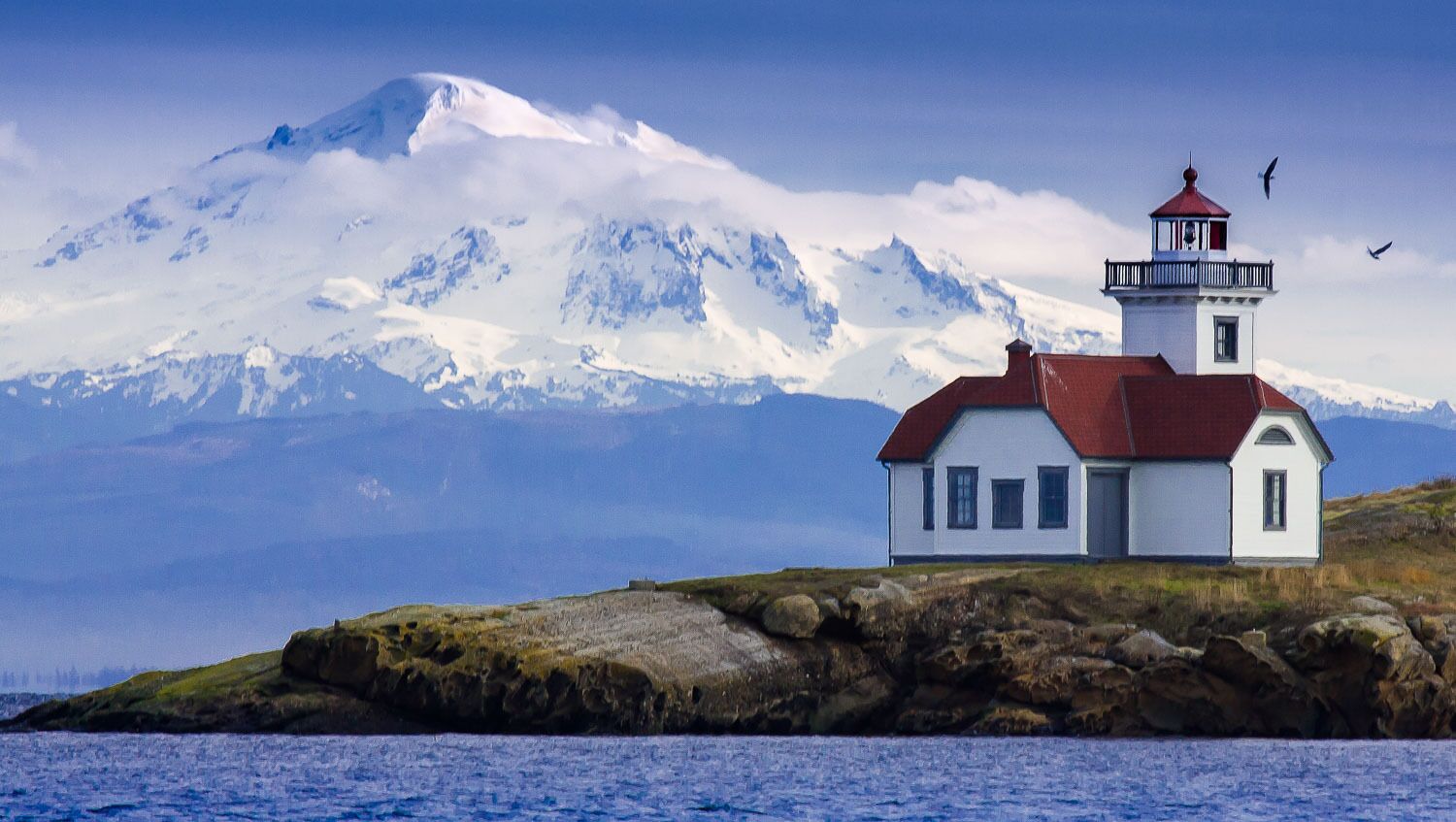 Un phare sur une île rocheuse, avec une montagne enneigée en arrière-plan.