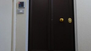Uma porta de madeira escura com puxador de latão e olho de fechadura, e um sistema de interfone fixado na parede à esquerda.