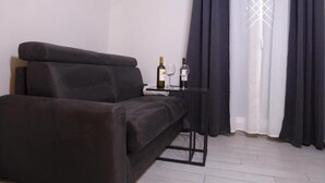 Uma sala de estar moderna com um sofá escuro, uma mesinha com garrafa e copo, e uma janela com cortinas escuras.