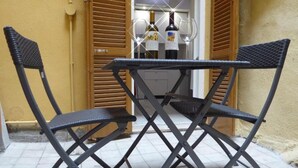 Uma pequena área externa com assento, com duas cadeiras dobráveis e uma mesa, posicionada em frente a uma porta com persianas de madeira.