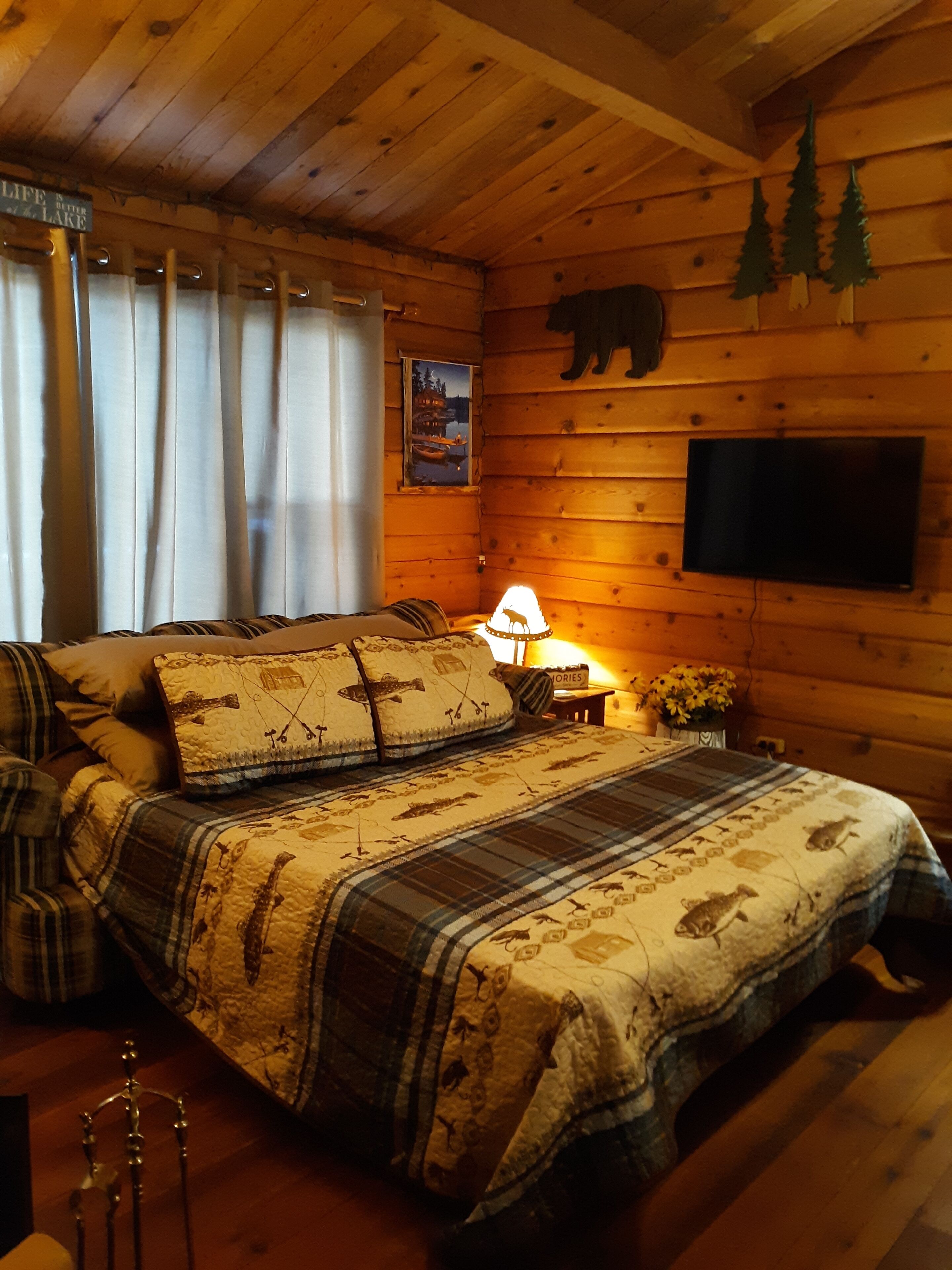 Une chambre confortable, aménagée dans un chalet en bois, avec un lit, des rideaux, une lampe, un téléviseur et une décoration représentant un ours sur le mur.