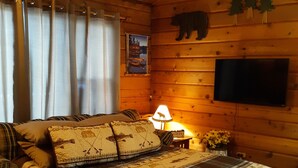 A cozy wooden cabin bedroom with a bed, curtains, a lamp, a TV, and a bear decoration on the wall.
