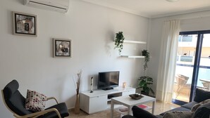 A modern living room with a flat-screen TV, a sofa, a rocking chair, and a coffee table. There are two potted plants, one on a shelf and one on the floor. - To Rent - Stunning 2 Bed Apartment in Cabo Roig, Spain (Dehesa de Campoamor)