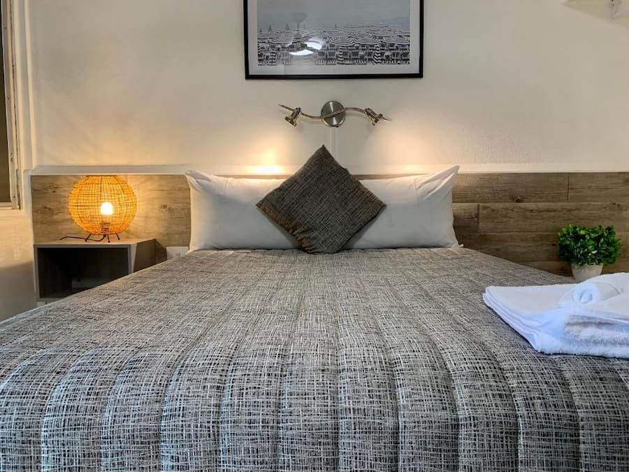 Een net opgemaakt bed met een gebreid plaid, een bedlampje, een ingelijst foto op de muur en een kleine plant in een pot.