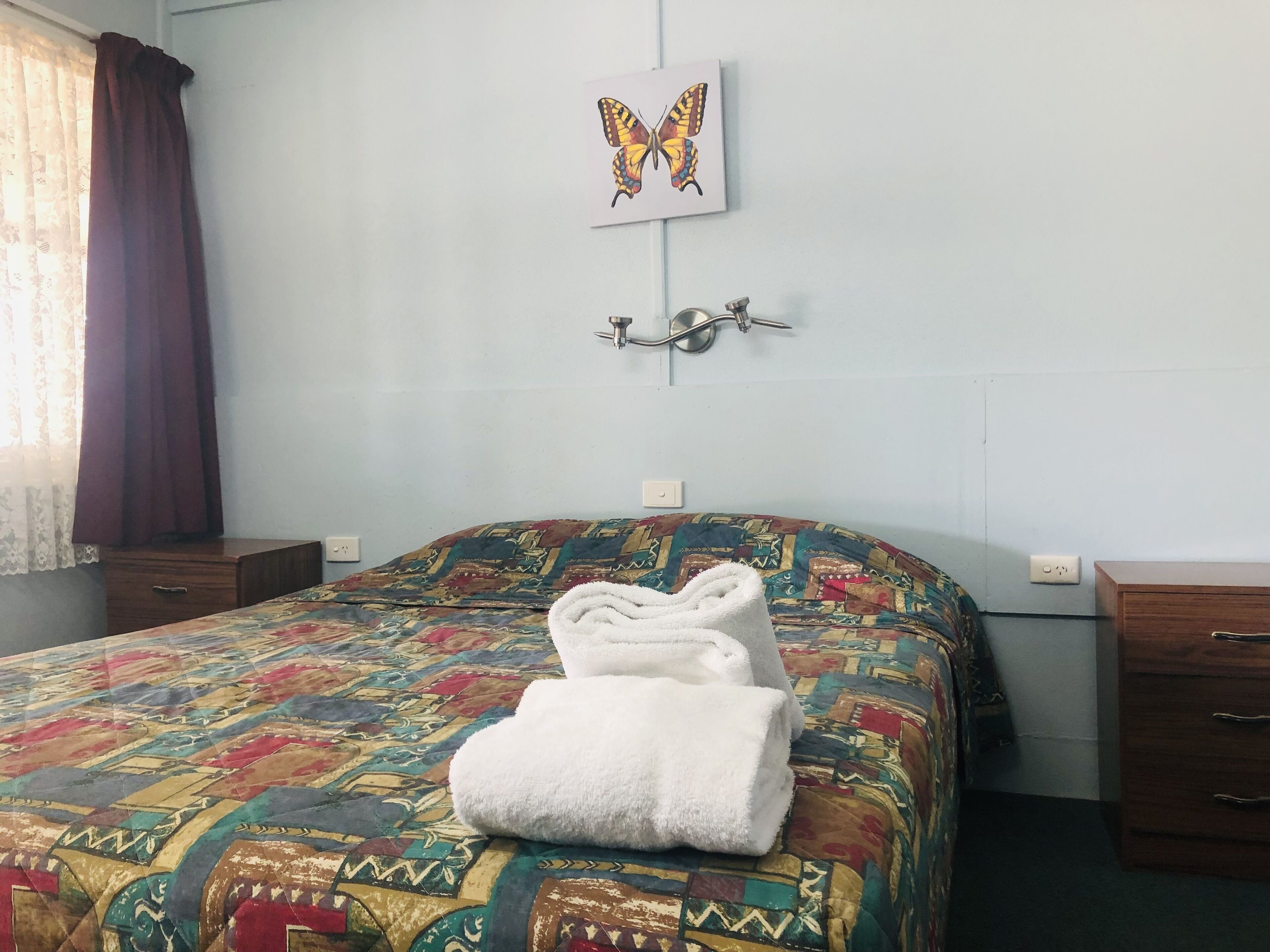 A neatly made bed with a colorful patterned bedspread, a folded white towel on the bed, a wall-mounted light fixture, and a framed butterfly artwork above the bed.