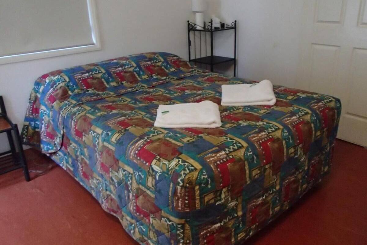 Ein Bett mit farbenfroher Steppdecke, ein Nachttisch mit Lampe und ein Fenster mit Jalousien.