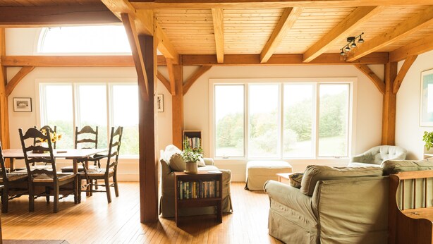 Amplia sala de estar con piso de madera, un comedor con sillas de madera, un sofá y una estantería. Amplias ventanas ofrecen luz natural y vistas al exterior.