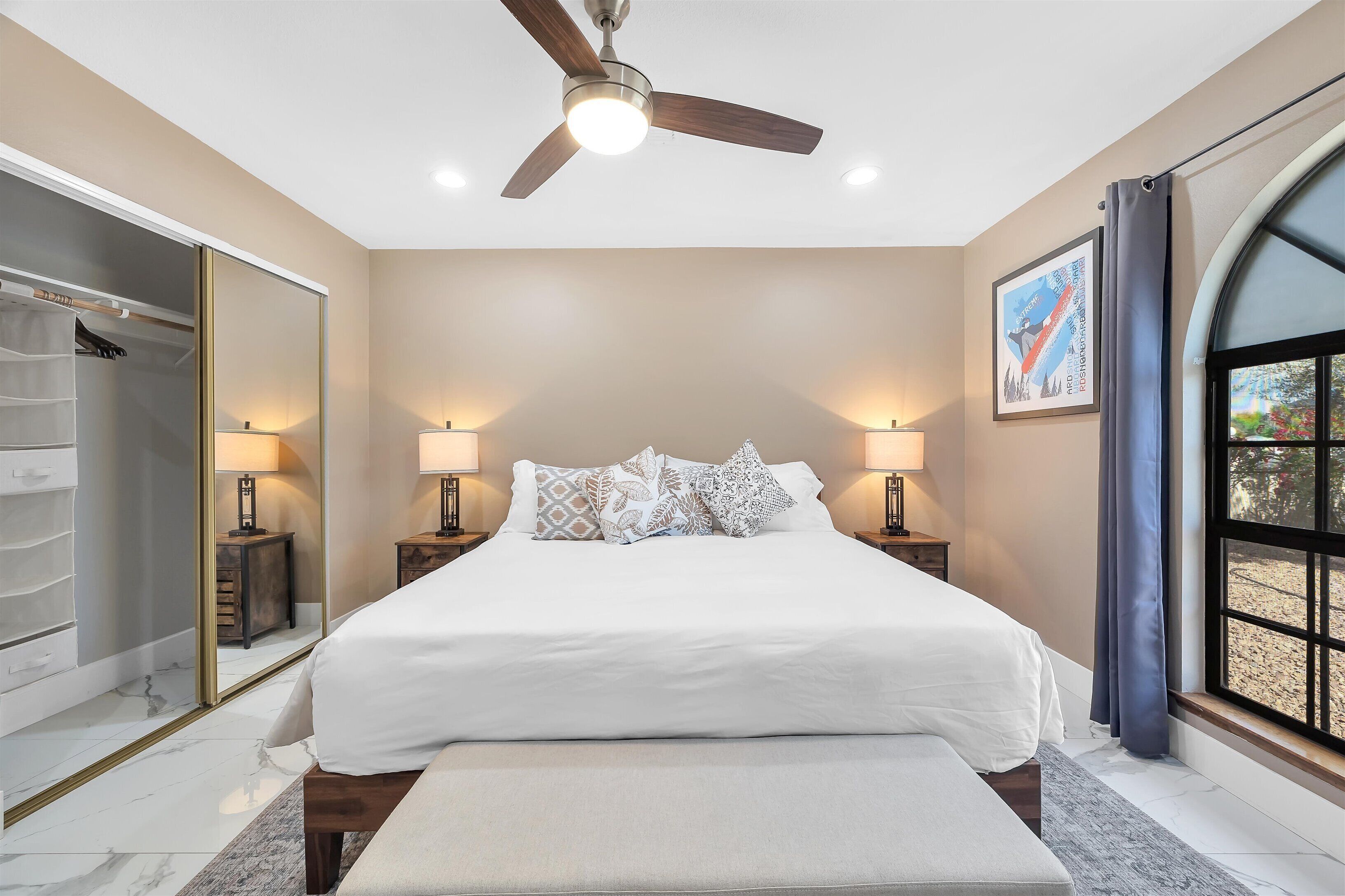A bedroom with a large bed, bedside lamps, a ceiling fan, a wardrobe, and a window with curtains.