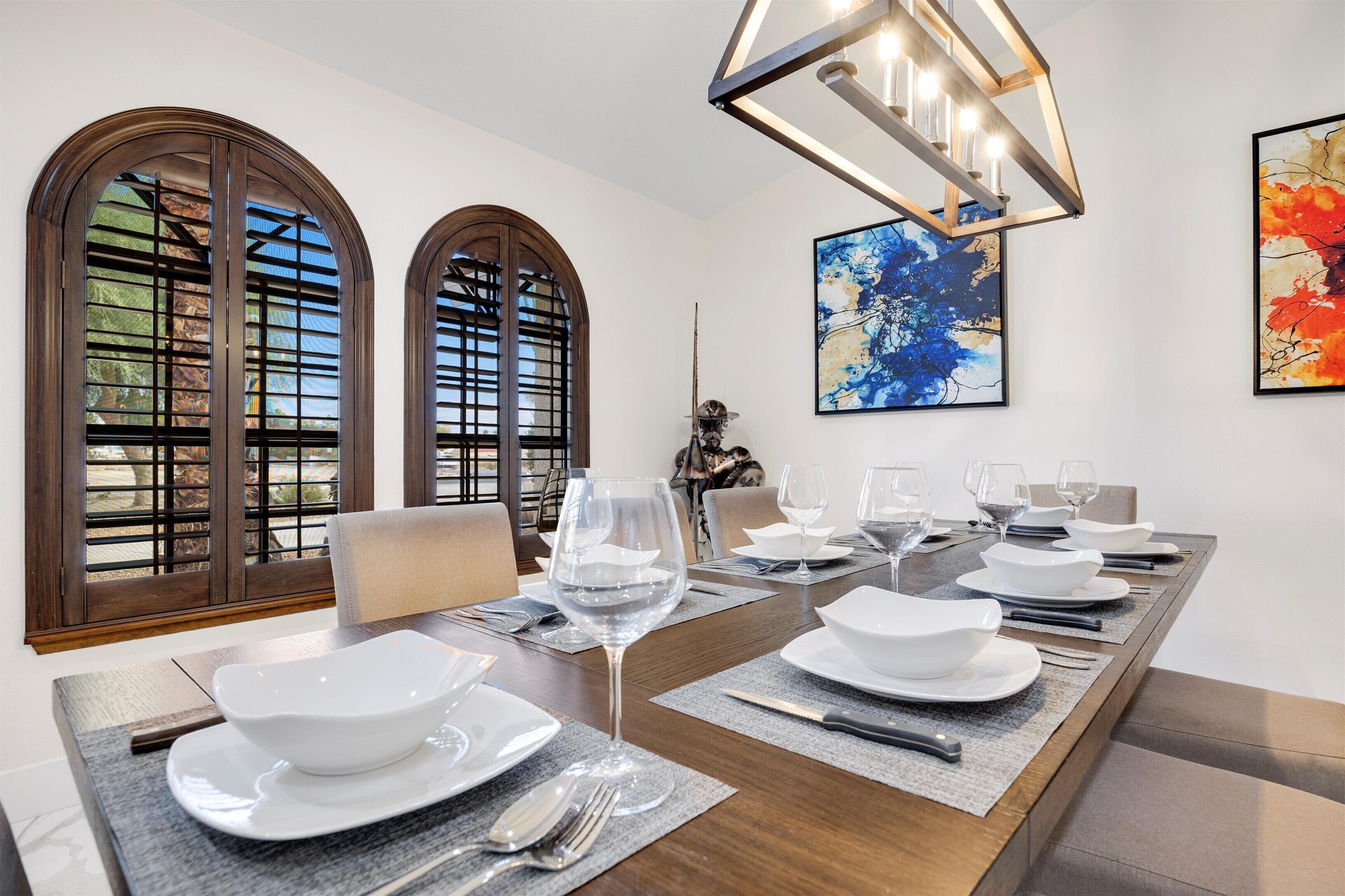 A dining room with a long table set for six, large arched windows, and a modern chandelier.