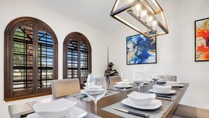 Een eetkamer met een lange tafel voor zes personen, grote gewelfde ramen en een moderne kroonluchter.