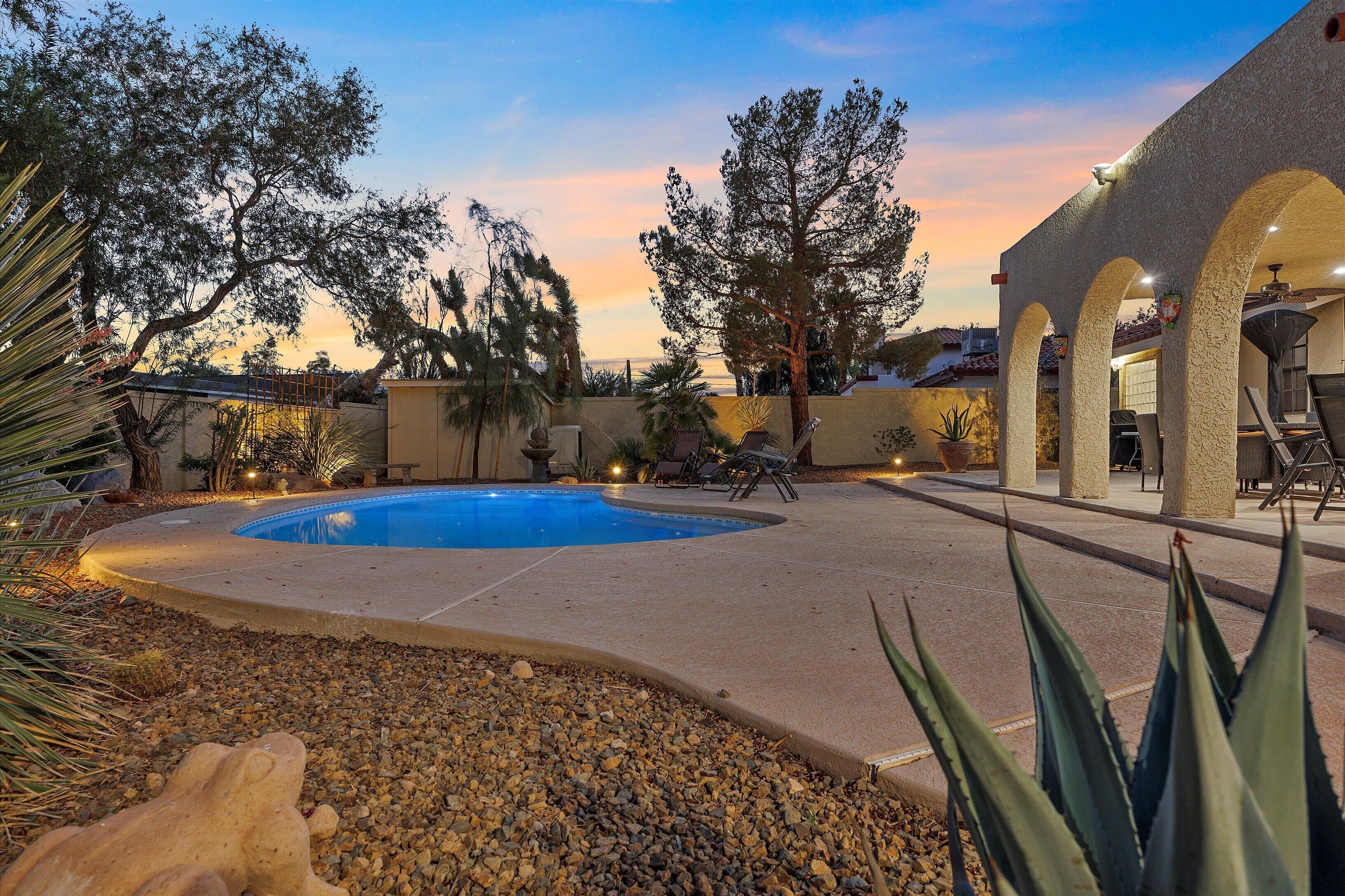 A pool area with a curved edge, surrounded by a patio with chairs and a table, and a building with arches in the background.